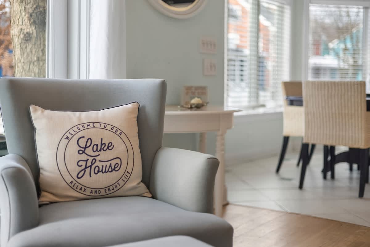 A close-up of a light gray armchair with a "Welcome to our Lake House" pillow, positioned near a window with white blinds, offering a glimpse of a dining area in the background.