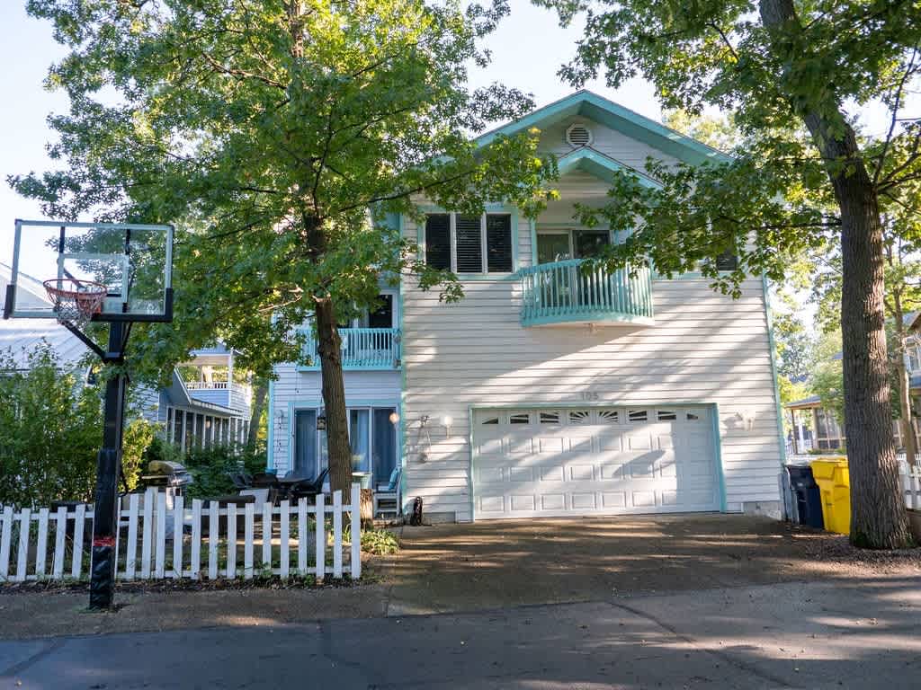 Front exterior of a white house with light blue trim, featuring a two-car garage, a second-story balcony, and a basketball hoop in the paved driveway, surrounded by trees.