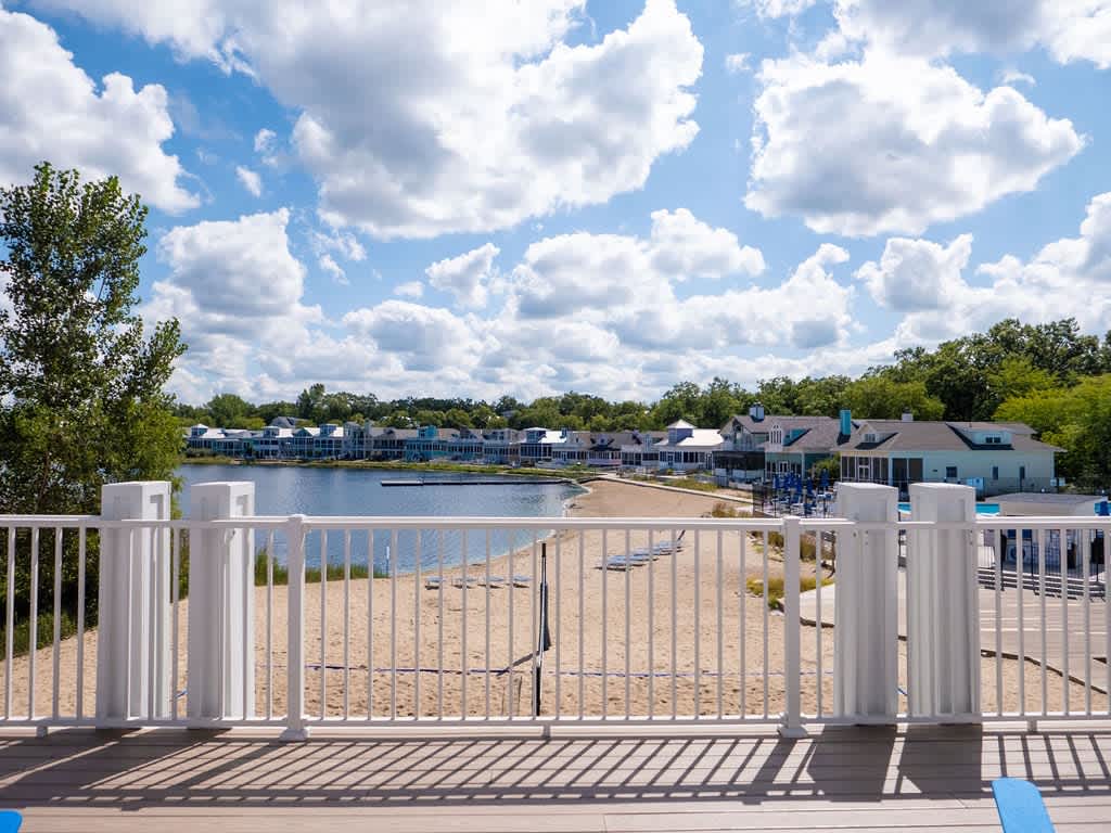 A scenic view from a wooden deck overlooks a wide sandy beach leading to a calm lake, with numerous houses lining the distant shore under a bright blue sky with fluffy white clouds.