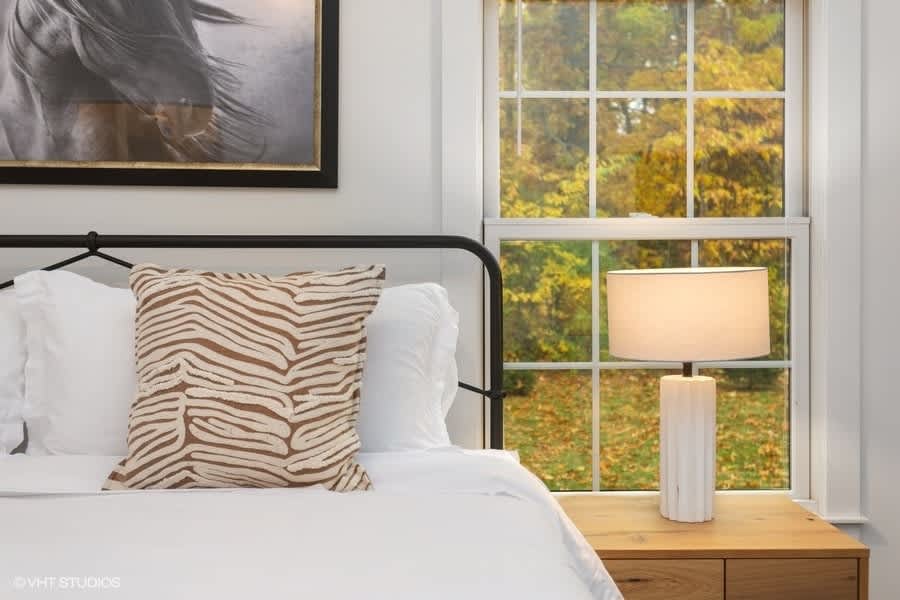 A cozy bedroom features a bed with white linens and a black metal headboard, accented by a brown and white patterned pillow. A modern white lamp sits on a light wood nightstand beside a large window overlooking vibrant autumn foliage.