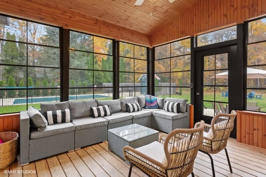 Screened-in porch with light wood paneled walls and ceiling, featuring large black-framed windows overlooking a wooded backyard and pool. A gray sectional sofa and wicker chairs provide seating.