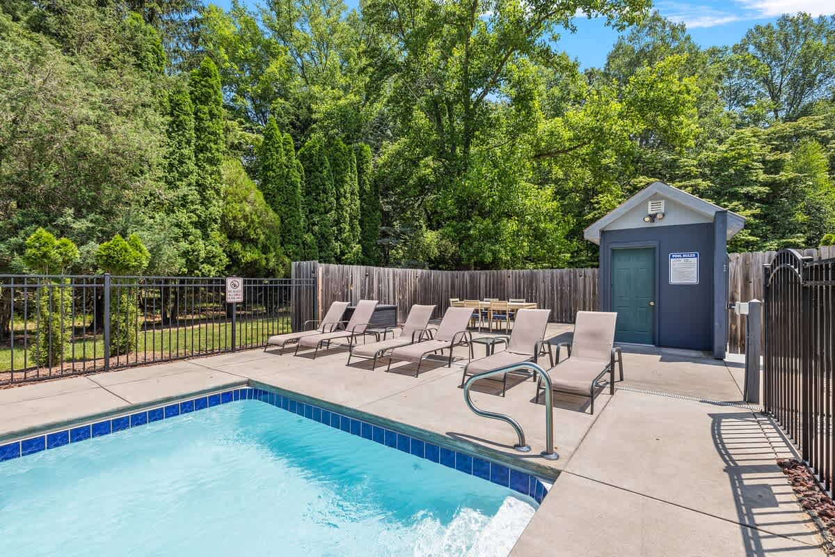 This view of the outdoor pool area highlights several lounge chairs arranged on the concrete deck, alongside a small pool house or shed. The entire space is enclosed by a black fence and surrounded by dense green foliage.