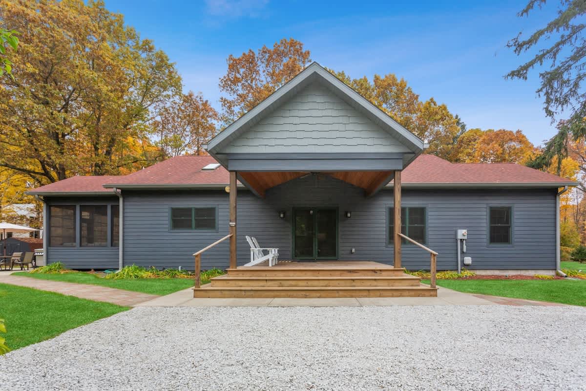 Modern single-story house with dark gray siding and a prominent covered front porch with wooden steps. The surrounding yard features gravel and green grass, with mature trees displaying autumn colors.