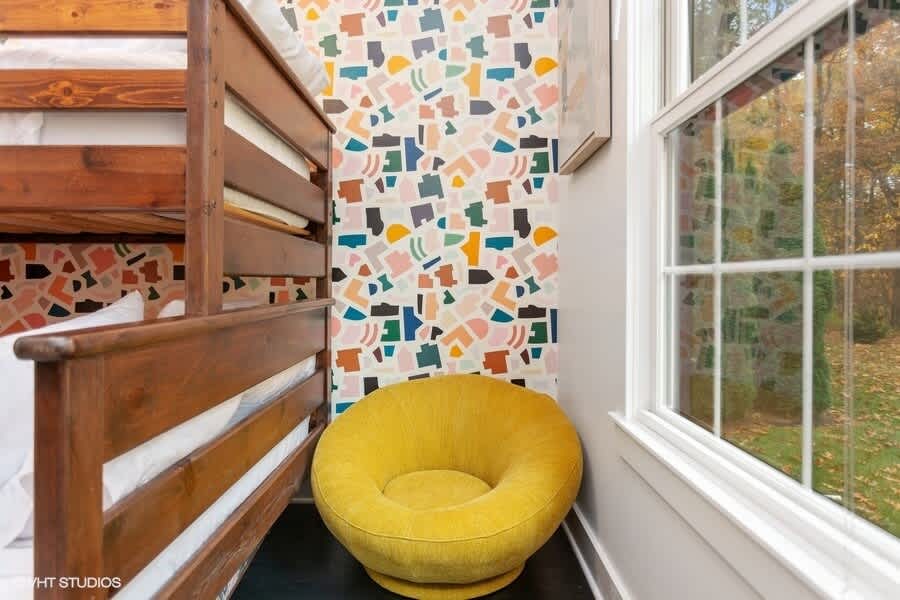A corner of the children's bunk room showcases the colorful abstract wallpaper and the sturdy wooden bunk bed frame. A bright yellow, plush round chair is tucked neatly beside a window, offering a cozy reading nook.