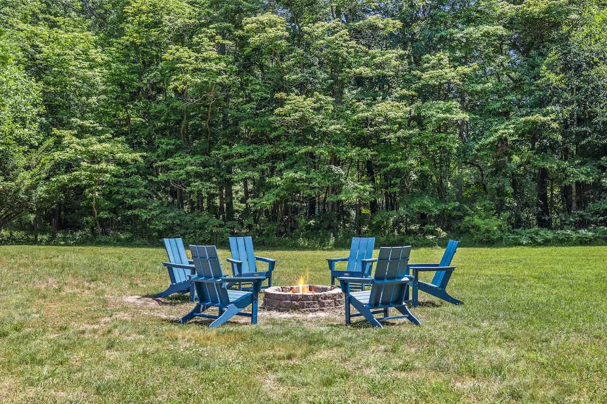 A circular stone fire pit is centrally placed in a lush green lawn, encircled by six blue Adirondack chairs, with a dense backdrop of tall green trees under a bright sky.