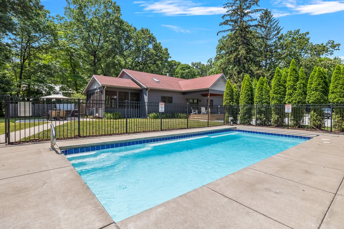 A sparkling rectangular outdoor pool is surrounded by a concrete deck and a black metal fence, with lounge chairs visible. The main house is in the background, nestled among tall green trees under a bright blue sky.