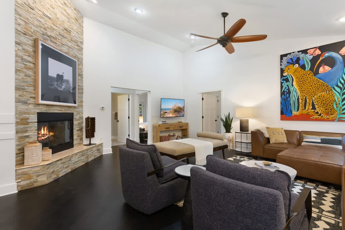 Spacious living room with a stone-clad fireplace, dark hardwood floors, and a brown leather sectional. A large, colorful cheetah artwork hangs above the sofa, complementing the bright white walls.
