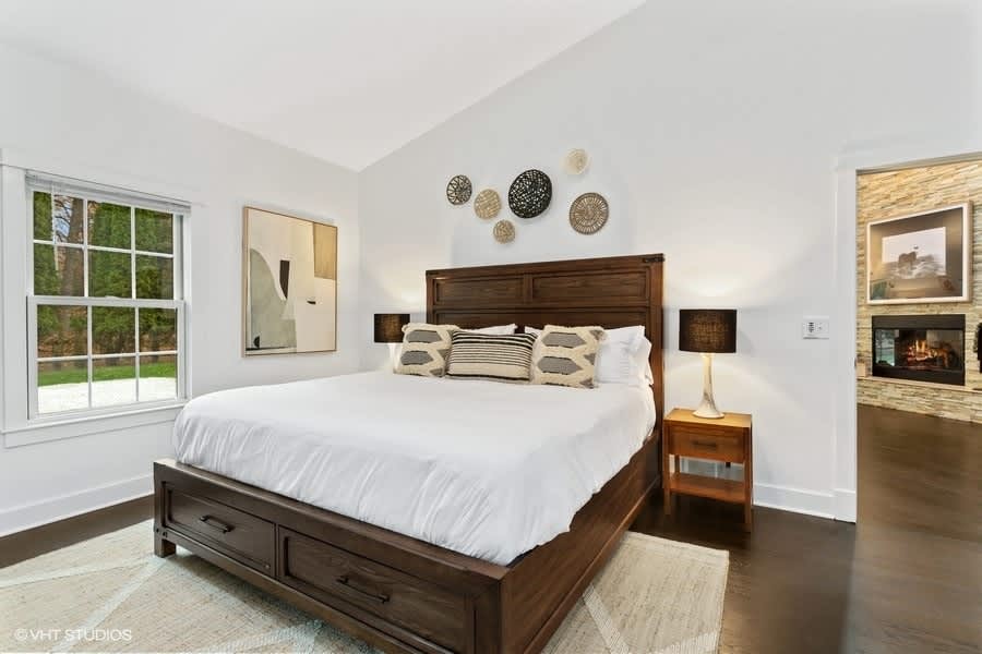 Serene bedroom with a dark wood king-sized bed, white bedding, and light-colored rug on dark hardwood floors, accented by circular wall art and a large abstract painting, with a glimpse of a stone fireplace beyond.