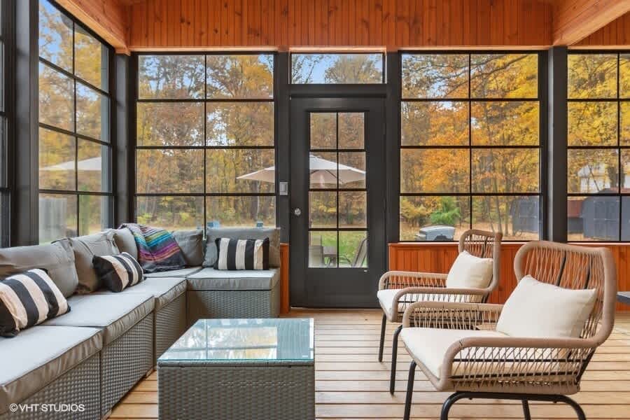 Spacious enclosed porch with dark-framed windows showcasing autumn foliage, featuring a gray sectional sofa with striped pillows, two wicker chairs, and a glass-top coffee table on a light wood floor.