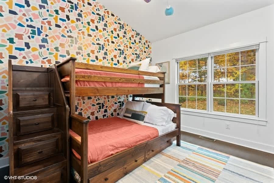 A vibrant children's bedroom features sturdy wooden bunk beds with coral bedding against a wall adorned with colorful, abstract patterned wallpaper. A large window offers views of autumn trees, and a striped rug adds to the playful atmosphere.