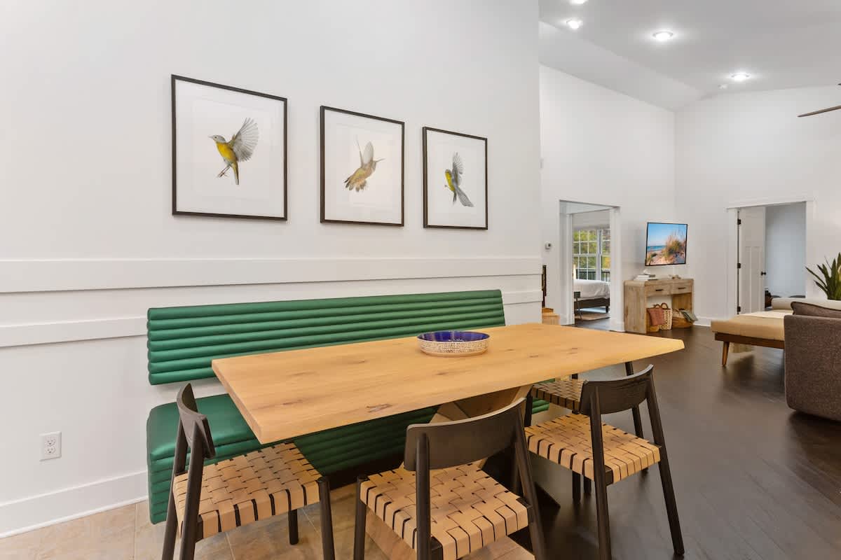Dining area featuring a long wooden table with a built-in green upholstered bench and modern chairs. Three framed bird artworks adorn the white wall above the bench, with the living room visible in the background.