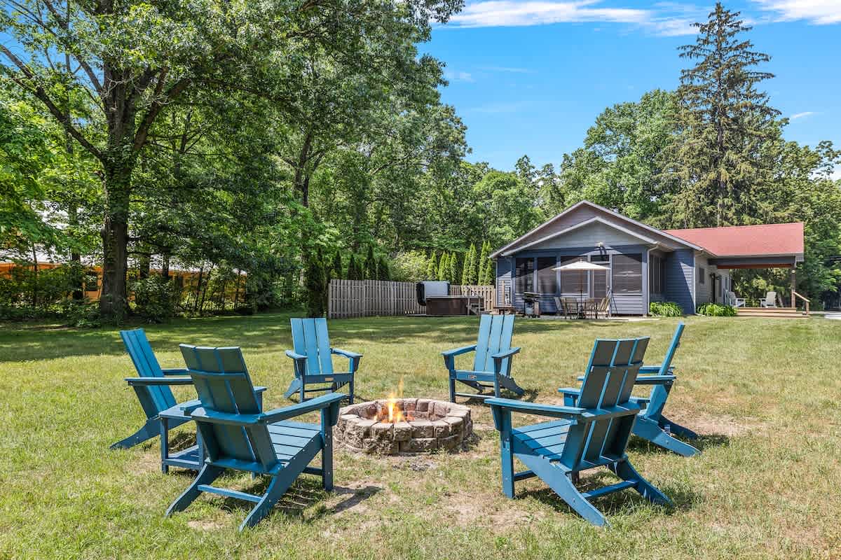 A inviting outdoor fire pit area features a circle of blue Adirondack chairs surrounding a stone fire pit, set on a large, well-maintained grassy lawn. The main house is visible in the background, framed by mature trees.