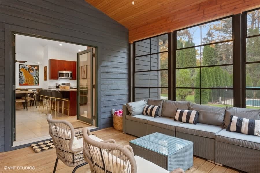 Enclosed porch with dark gray siding and large black-framed windows, furnished with a gray sectional sofa and two wicker chairs, offering a view into the kitchen through open French doors.