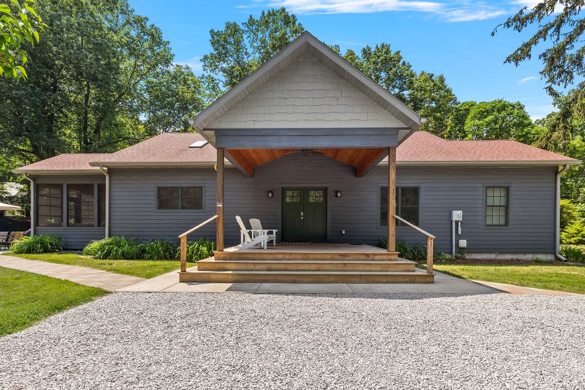 The front exterior of a modern single-story house features gray siding, a large covered porch with a light gray gable roof, and a wide gravel driveway leading to the entrance, flanked by green landscaping.