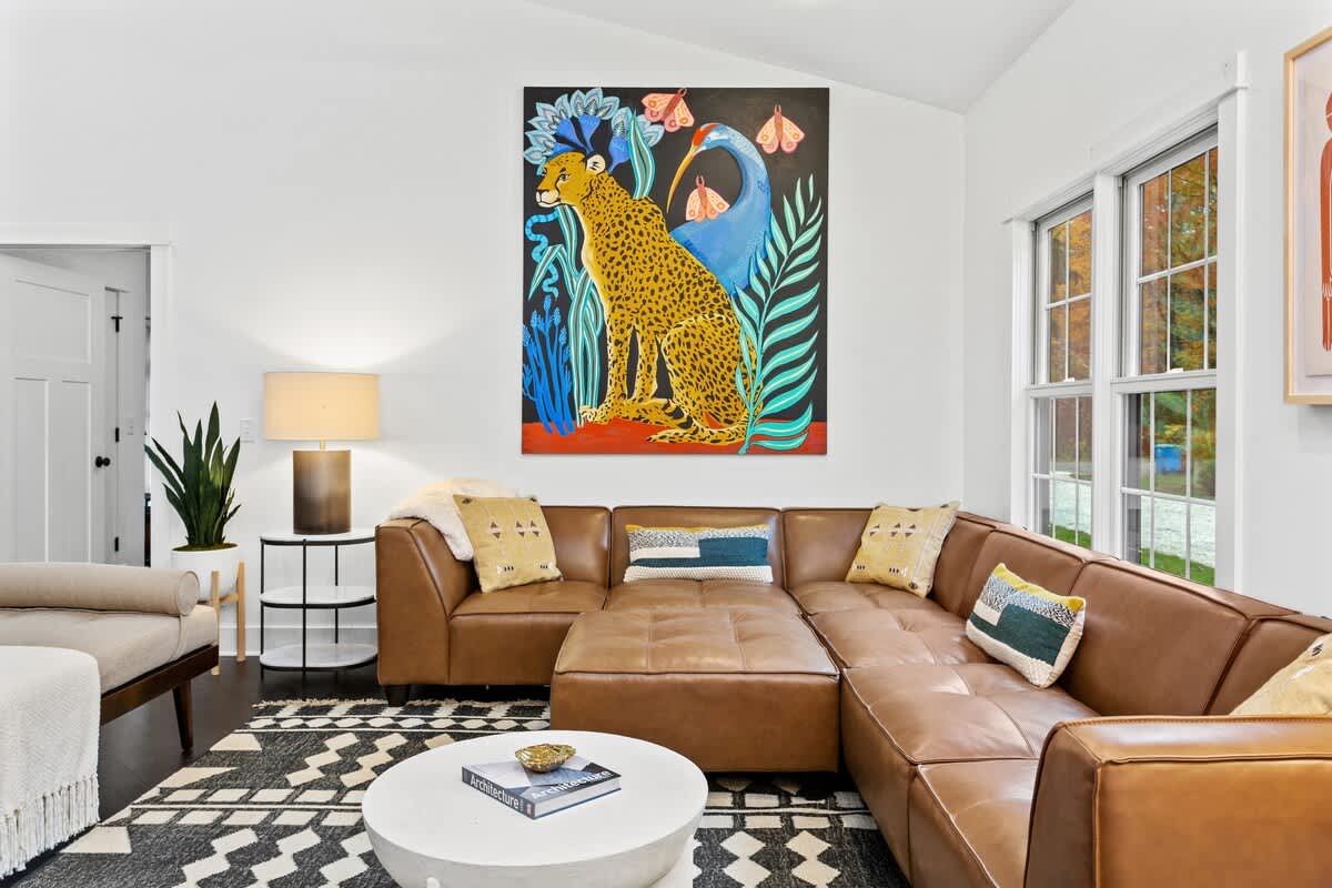 Cozy living area featuring a brown leather sectional sofa adorned with yellow and green throw pillows, set on a black and white geometric rug. A vibrant cheetah painting dominates the white wall.