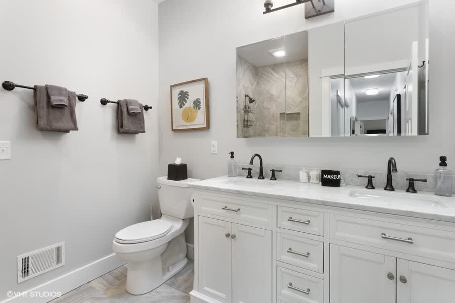 A modern bathroom features a double vanity with white shaker cabinets, a marble countertop, and two rectangular sinks with black faucets. A large mirror reflects a tiled shower, while light gray walls and a toilet complete the space.