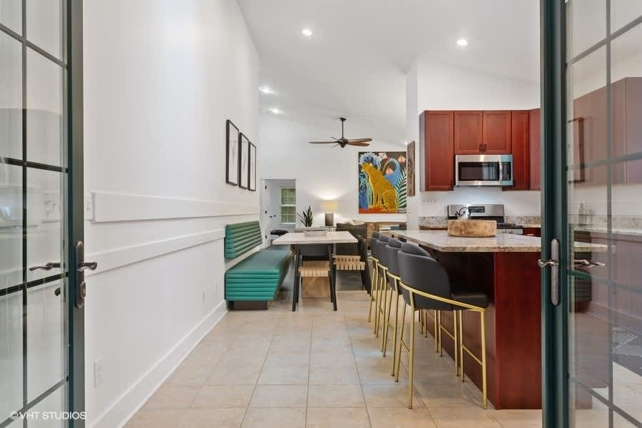 Open-concept kitchen and dining area with dark wood cabinets, granite island, and a teal banquette, leading to a living space with a colorful abstract painting, all under a vaulted ceiling with recessed lighting.