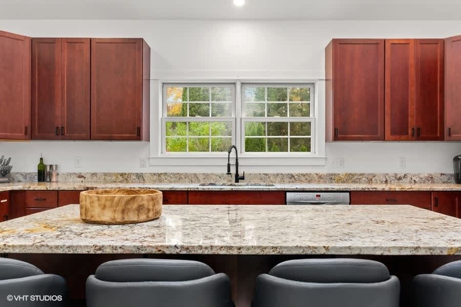 Bright kitchen featuring dark wood cabinetry, light granite countertops, a large island with seating, and a window overlooking lush greenery, with a black faucet and a wooden bowl as a centerpiece.