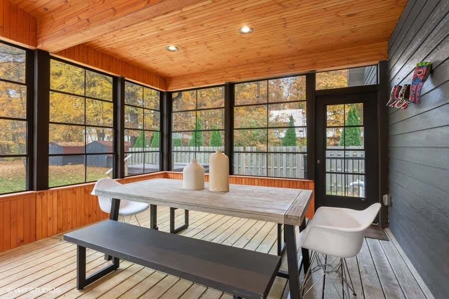 Bright enclosed porch with wood-paneled ceiling and floor, featuring a light wood dining table with a dark bench and two white chairs, surrounded by large windows offering views of the autumn landscape.