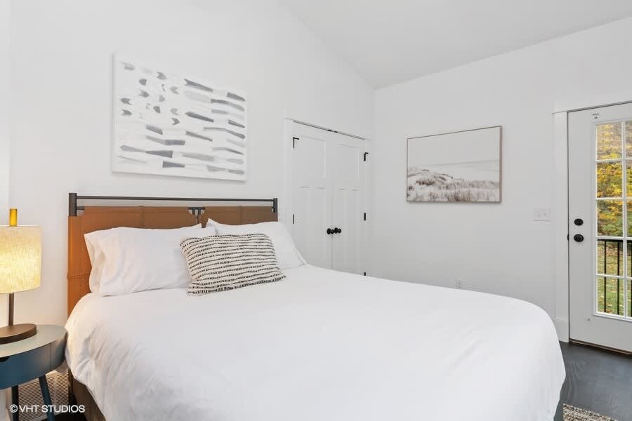 A bright bedroom with white walls and dark wood floors features a bed with white linens and a brown upholstered headboard. A black and white patterned pillow adds contrast, while a blue round side table with a lamp and two abstract art pieces complete the minimalist decor.
