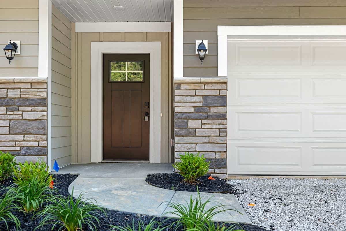 Front exterior of the house featuring a dark wood front door with a window, a white two-car garage, stone accents, and manicured landscaping with small bushes.