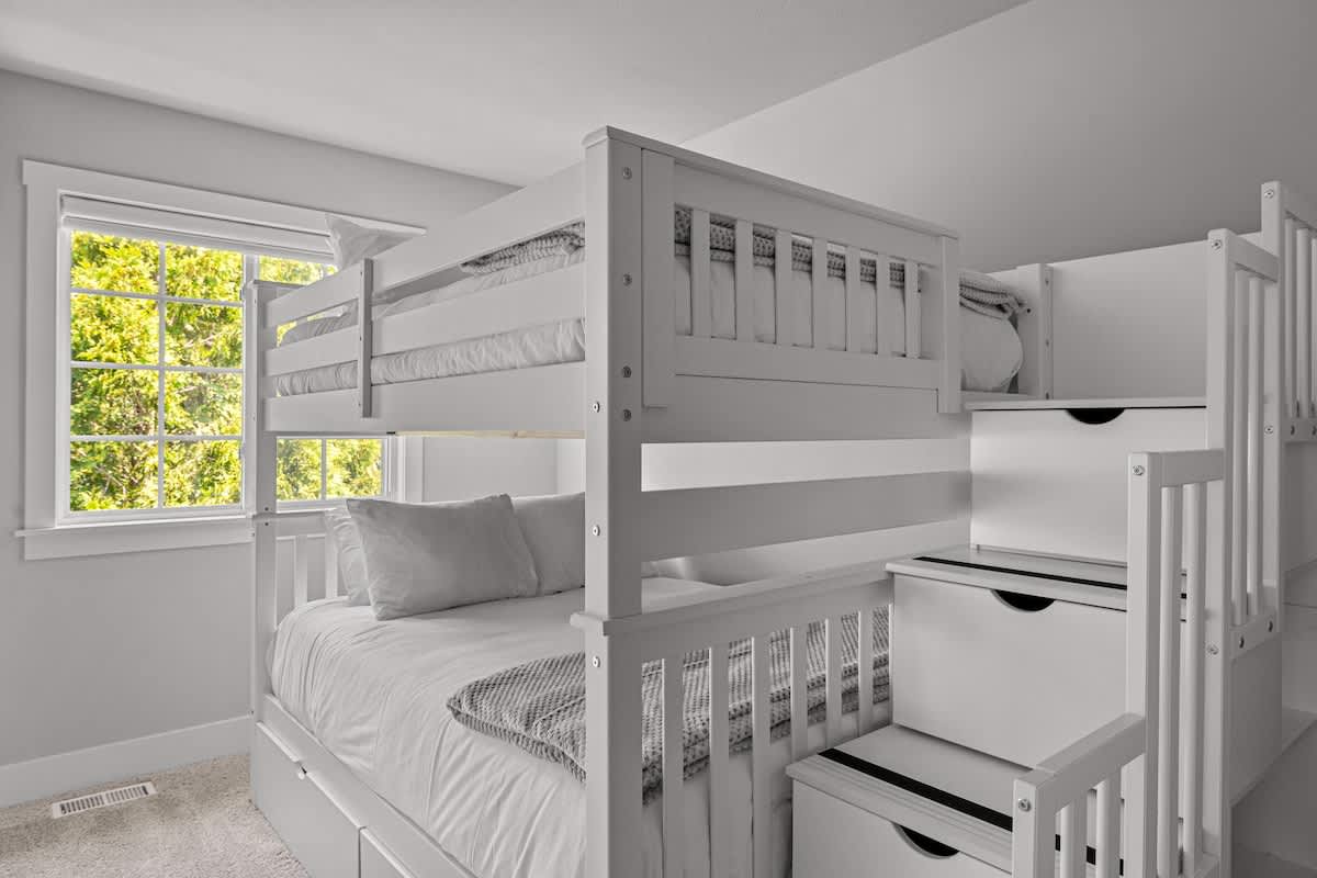 Children's bedroom with white wooden bunk beds, including a full bed on the bottom and a twin on top, complemented by built-in storage stairs and a window overlooking lush greenery.