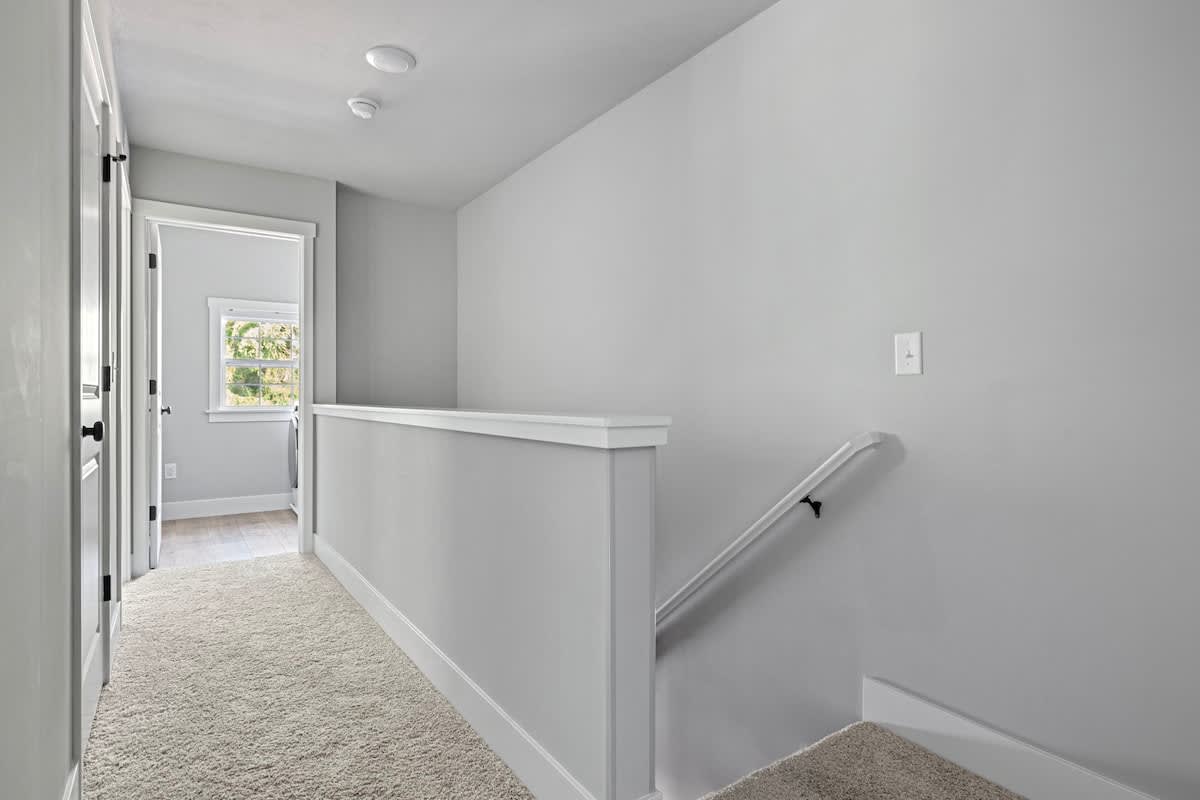 Upper-level hallway or landing with light gray walls, a carpeted floor, a white railing, and a glimpse of a room through an open doorway at the end.