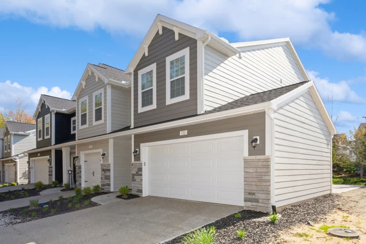 A row of modern townhouses with light gray and dark gray siding, white trim, and attached garages with white doors under a bright blue sky. The driveways are concrete, and small landscaping beds are visible.