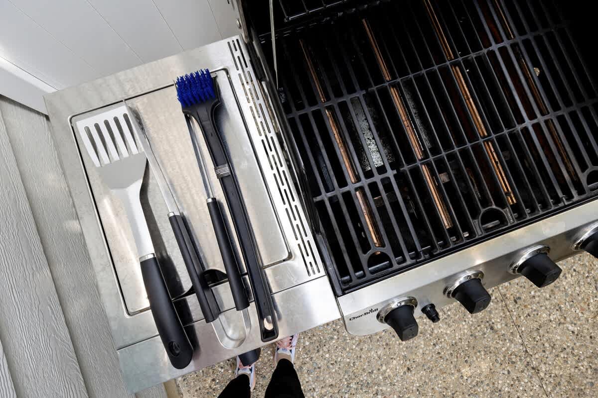 A close-up view of a stainless steel BBQ grill with its grates visible, accompanied by grilling tools including a spatula, tongs, and a brush, resting on the side shelf.