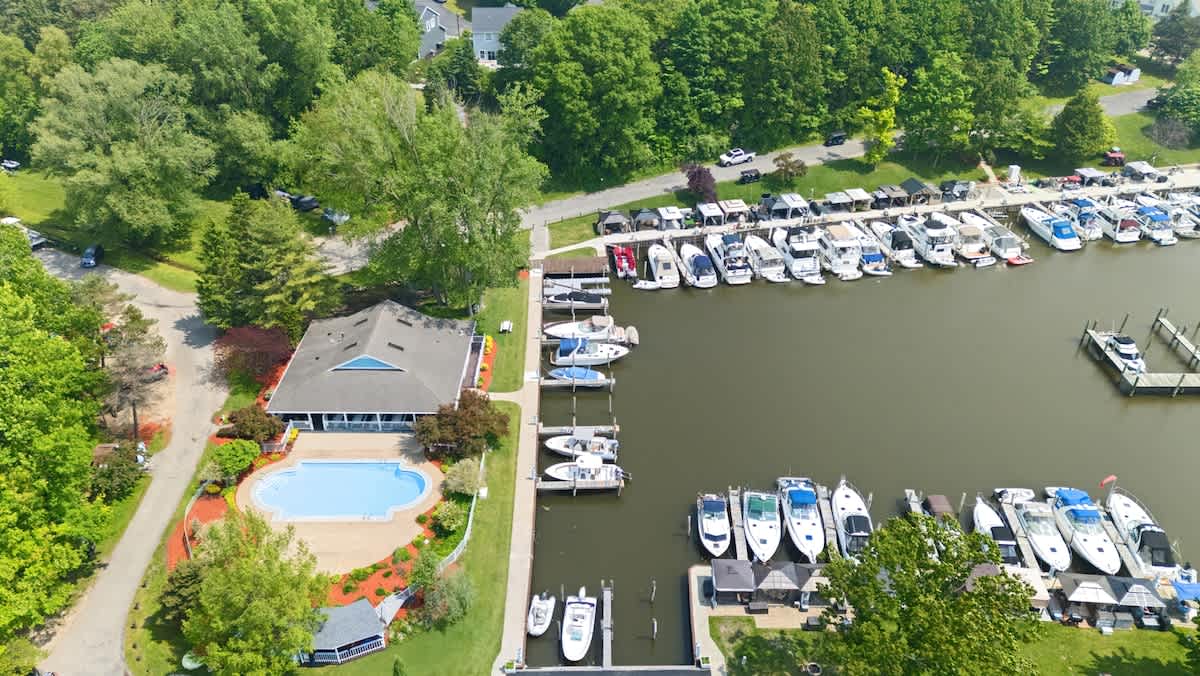 Aerial view of a bustling marina filled with numerous boats docked along a waterway, adjacent to a building with a swimming pool, all surrounded by dense green foliage and trees.