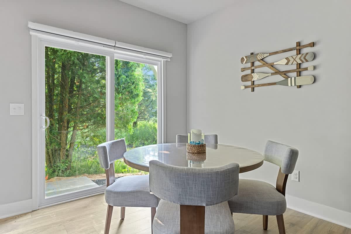 Dining area with a round table and four upholstered chairs, featuring a large sliding glass door that opens to an outdoor area with lush green trees.