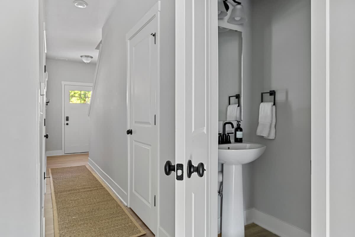 A bright hallway with light gray walls and a jute runner rug leads to a powder room featuring a white pedestal sink with a black faucet and mirror, and a white door with black hardware.