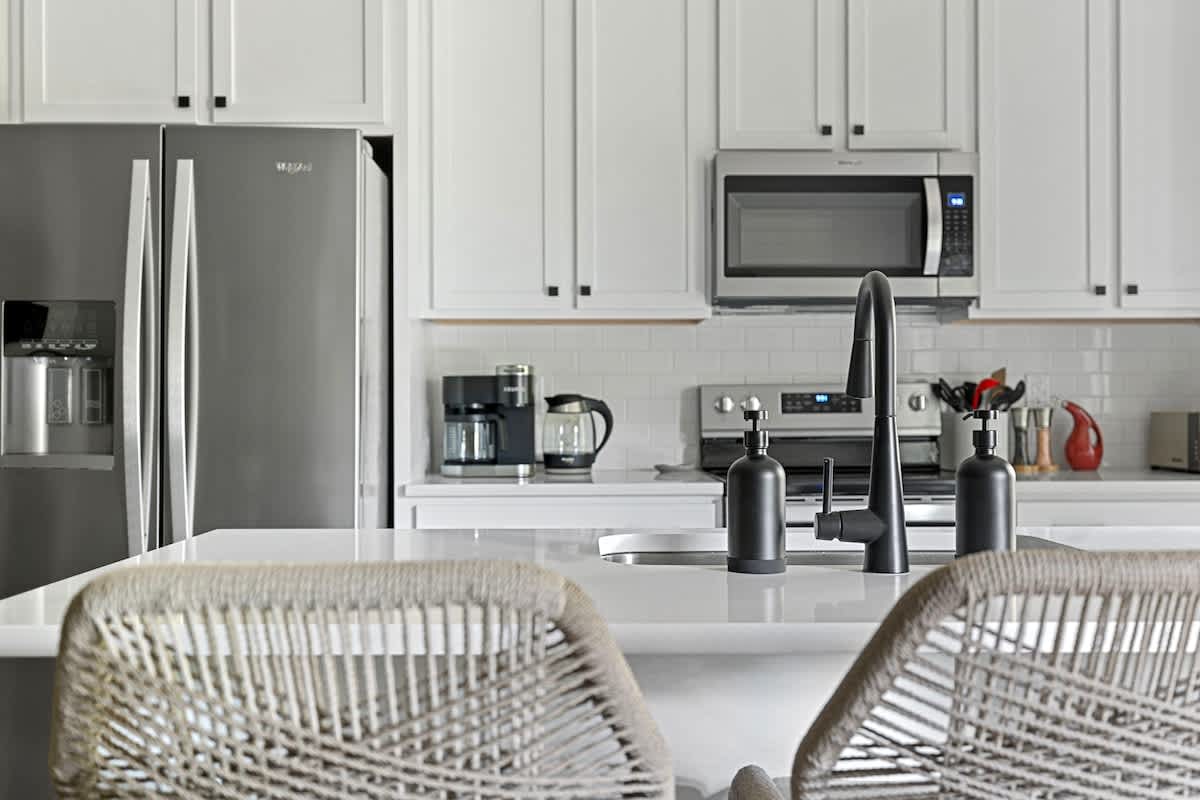 A modern kitchen with white shaker cabinets, stainless steel appliances including a refrigerator and microwave, and a white countertop island with a sleek black faucet. A coffee maker and other kitchen essentials are visible.