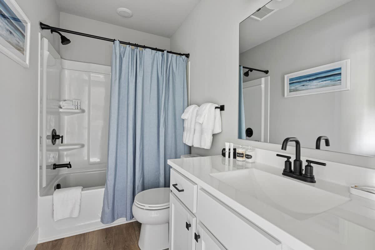 A modern bathroom features a white vanity with a single sink, black faucet, and large mirror, alongside a toilet and a shower/tub combo with a light blue shower curtain. The walls are light gray, and the floor is light wood-look vinyl.