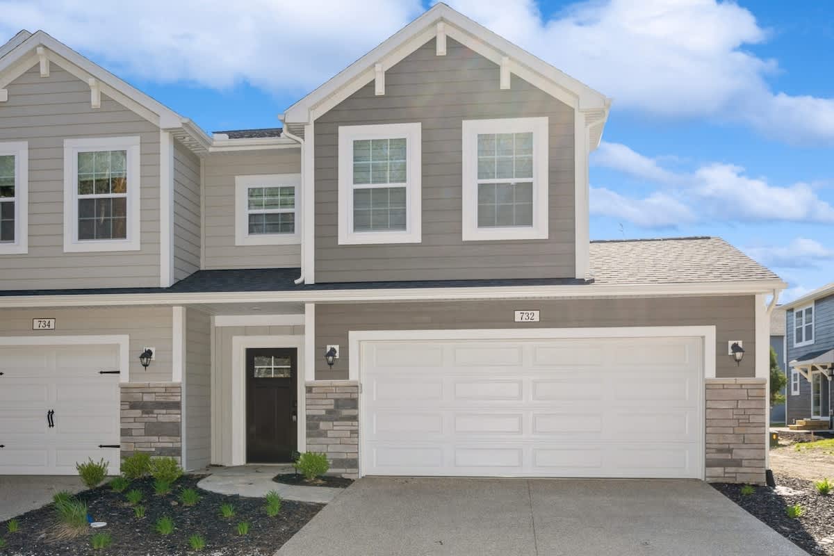 The front exterior of a modern townhouse with gray siding, white trim, and stone accents, featuring a two-car garage and a dark front door. The sky is blue with some clouds.