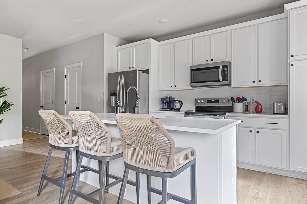 Bright kitchen featuring white shaker cabinets, stainless steel appliances including a refrigerator and microwave, and a large island with three woven bar stools.