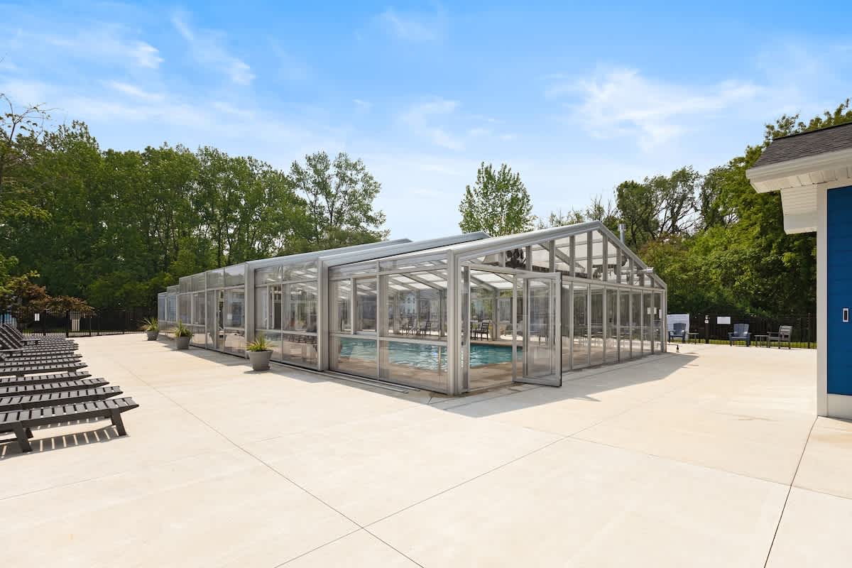 Wide shot of a modern, glass-enclosed swimming pool facility with a light concrete deck, surrounded by green trees and several dark lounge chairs, under a bright blue sky.