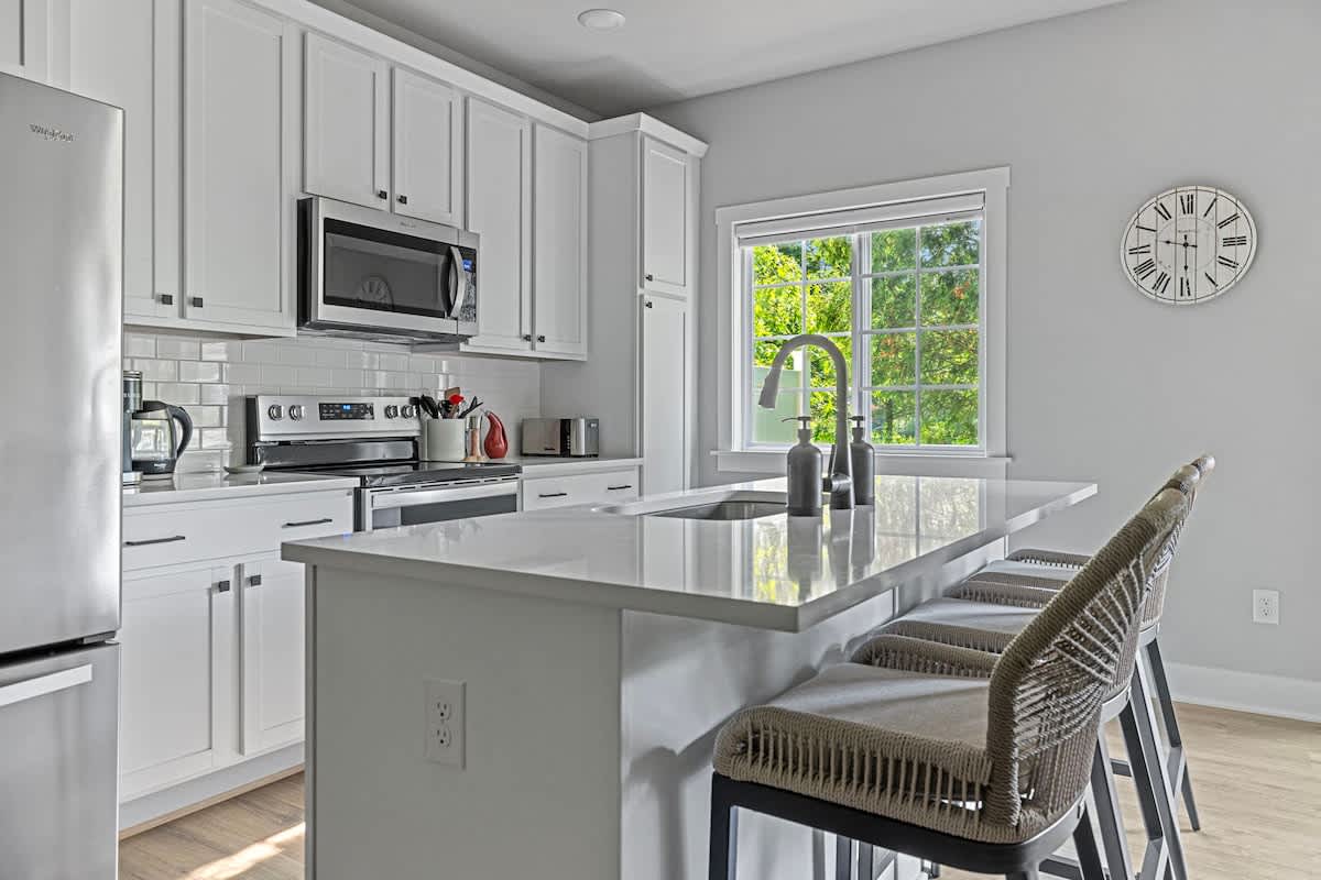Bright kitchen with white shaker cabinets, a stainless steel refrigerator, a microwave above the range, and a large island with bar seating, illuminated by natural light from a window.