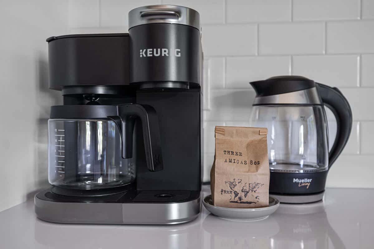 Close-up of a black Keurig coffee maker and a Mueller Living electric kettle on a white countertop, with a bag of coffee beans in between, against a white subway tile backsplash.