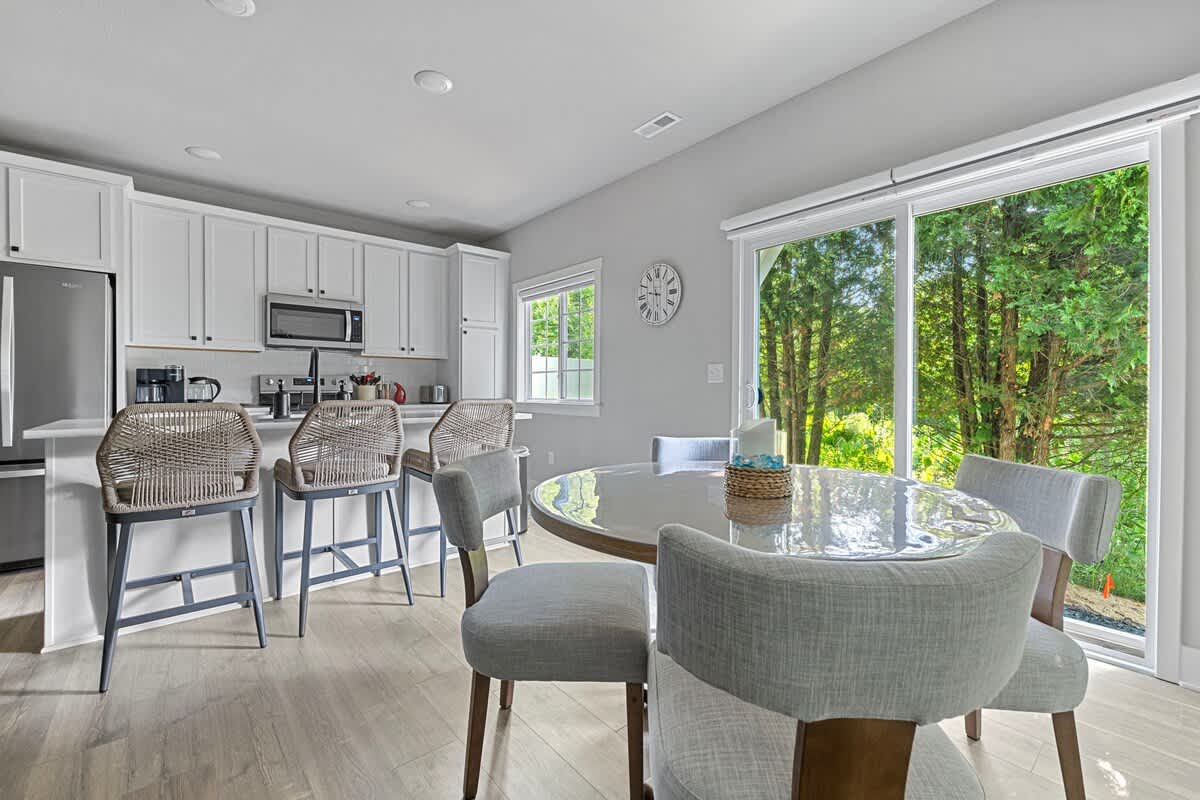 An open-concept kitchen and dining area featuring white cabinets, a kitchen island with bar stools, and a round dining table with four chairs. Large sliding glass doors lead to an outdoor area with trees.