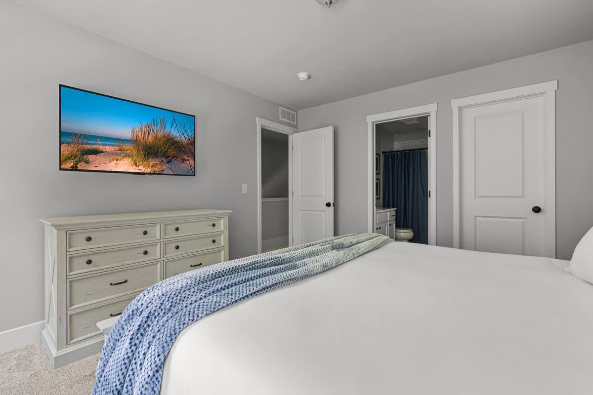 A spacious bedroom with light gray walls features a large bed with white bedding and a blue throw, a distressed white dresser, and a wall-mounted TV displaying a beach scene. An open door reveals a glimpse of an en-suite bathroom.