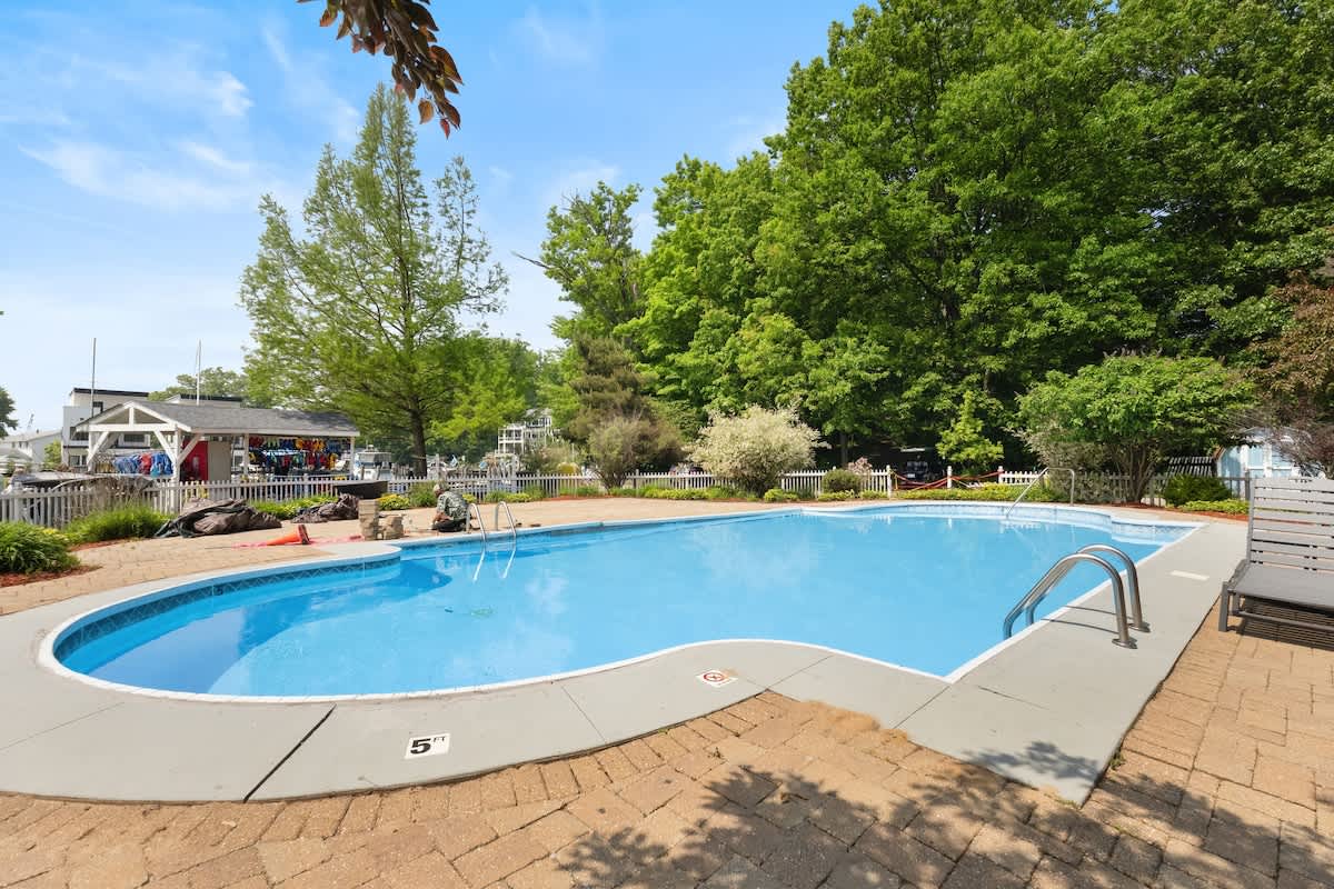 Outdoor swimming pool with a classic kidney shape, surrounded by brick paving and lush green trees, featuring a metal handrail for entry and a lounge chair on the side.