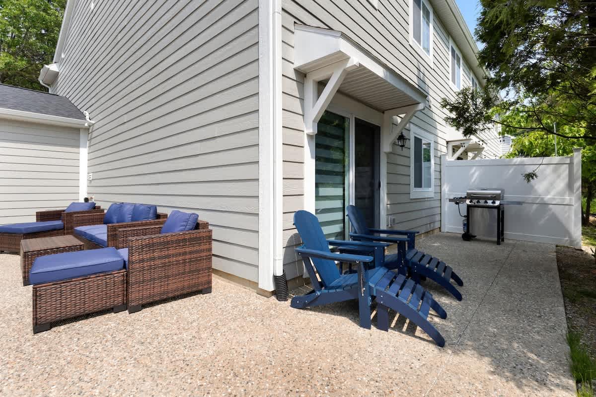 Outdoor patio area featuring light-colored concrete flooring, wicker-style seating with blue cushions, two blue Adirondack chairs, and a gas grill, all adjacent to a light-sided building.