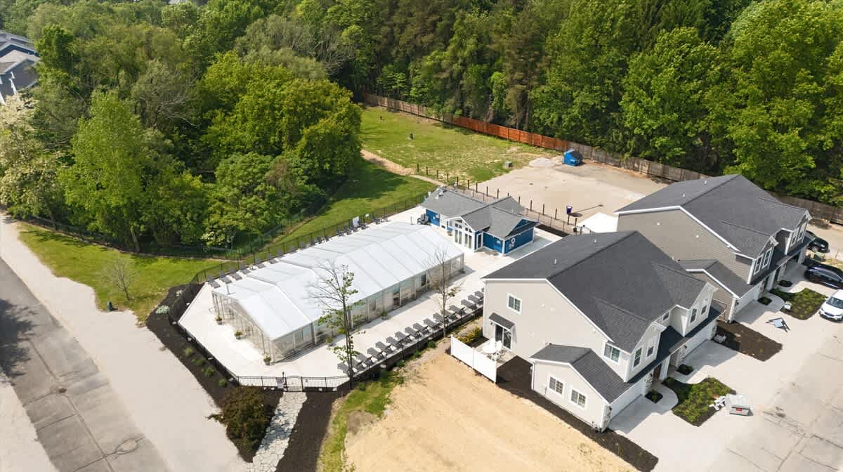 Aerial view showcasing a large, white tent-like structure, possibly a covered pool or event space, surrounded by a paved area and several modern residential buildings, all set amidst green trees.