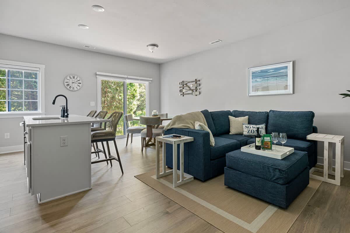 An open-plan living and dining area with a navy blue sectional sofa, a round dining table, and a kitchen island with bar seating. Light hardwood floors and light gray walls create a bright, modern space.