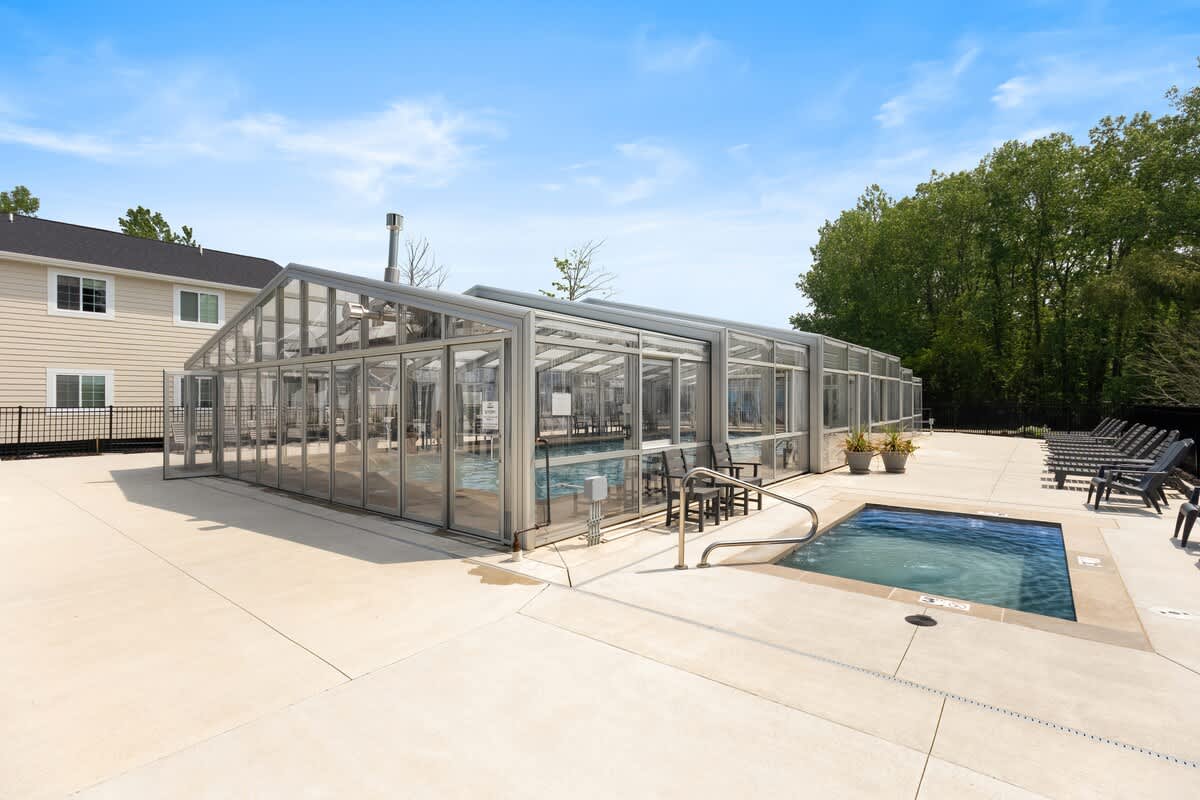 Expansive outdoor pool area with a large, glass-enclosed swimming pool structure and a separate, uncovered hot tub, surrounded by light concrete paving and several lounge chairs under a clear sky.