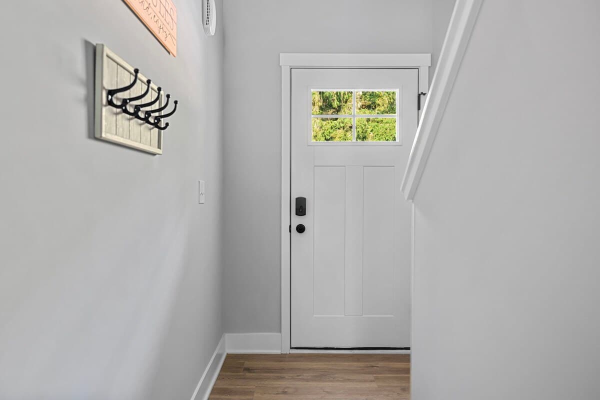 A welcoming foyer features a white front door with glass panes, a smart lock, and a coat rack with black hooks mounted on a light gray wall. The floor is light wood-look vinyl.