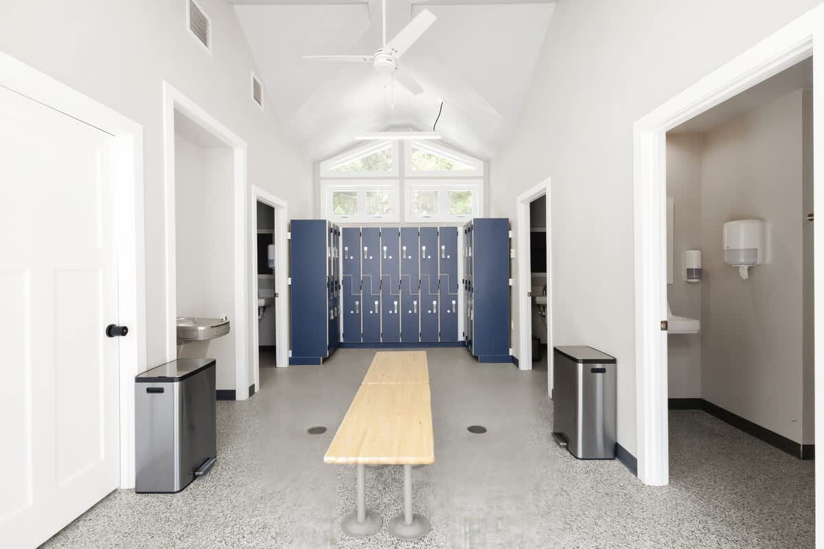 Bright, vaulted locker room with light gray speckled flooring, a long wooden bench, and two rows of dark blue lockers, featuring a large window overlooking trees and two stainless steel trash cans.