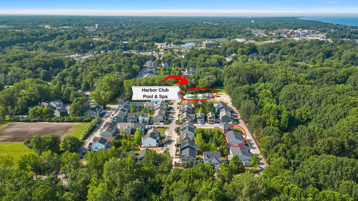 Aerial view of a residential community nestled among lush green trees, highlighting the 'Harbor Club Pool & Spa' area with a red arrow and circle, showing multiple houses and surrounding natural landscape.