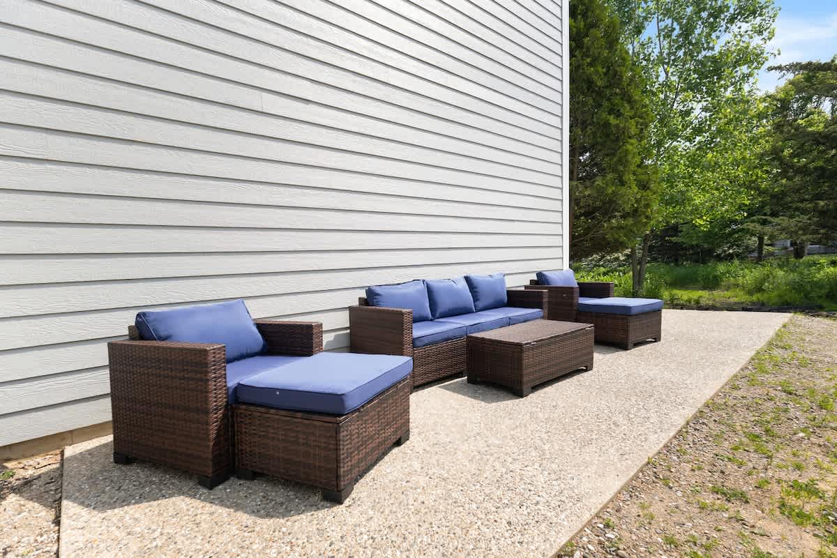A concrete patio area alongside the house, furnished with a brown wicker outdoor sofa, two armchairs, and an ottoman, all with blue cushions. Green trees and grass are visible in the background.